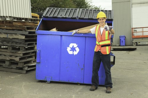A male waste collection worker wearing a yellow safety helmet, reflective orange and yellow vest, and gloves, standing next to a large blue commercial rubbish skip with an open top, revealing some discarded waste inside. The skip features a white recycling symbol on the front and is positioned on a paved surface in an industrial setting, with stacked pallets to the left and a large metal warehouse building in the background. The worker is smiling and giving a thumbs-up gesture, suggesting a waste removal or rubbish clearance operation typical of professional services in the Chiswick area of west London. The scene is well-lit with natural daylight, highlighting the textures of the skip's metal surface, the worker’s clothing, and surrounding environment. This imagery reflects the professional rubbish clearance services offered by Commercial Waste Chiswick within the local industrial and commercial areas, emphasizing proper waste management support in the vicinity.