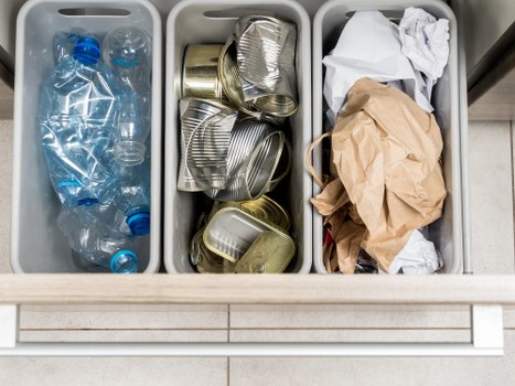 A close-up of three grey plastic rubbish sorting bins positioned in a commercial or industrial setting, likely outdoors or on a pavement, with a tiled surface beneath. The left bin contains several clear plastic bottles with blue caps, some crushed and partially flattened. The middle bin holds a collection of metal cans, including aluminium and tin, some stacked and others lying flat, with visible ridges and open tops. The right bin contains crumpled paper and brown paper bags, appearing flattened and loosely gathered, with some loose paper resting on the top. The scene emphasizes waste segregation and disposal, typical of commercial waste clearance services such as those offered by Commercial Waste Chiswick, serving the Chiswick area, with the environment suggesting an organized approach to rubbish removal near a business or retail location in West London.