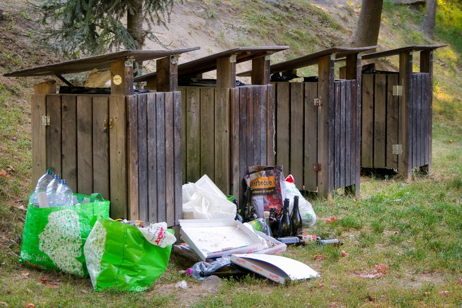 Three large black refuse bags filled with waste material are placed on the pavement beside a wooden fence with vertical slats. The bags appear to be made of thick plastic and are somewhat crumpled and